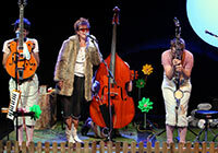 Drei Schauspieler mit Instrumenten auf einer Bühne mit großen Blumen