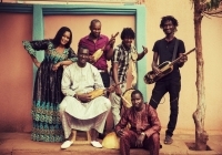 Eine Gruppe Männer und Frauen mit dunkler Hautfarbe und in afrikanischer Tracht steht vor einer einfachen Hauswand und lächelt mit Instrumenten in die Kamera.