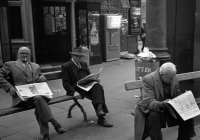 Das schwarz-weiß Bild von dre Männern, die auf Bänken sitzen und Zeitungen lesen. 