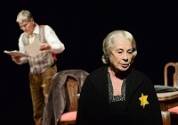 Ein Mann steht im Hintergrund und hält Papier in der Hand, das er liest. Im Vordergrund sitzt eine ältere Frau mit schwarzer Kleidung und gelbem Stern.
