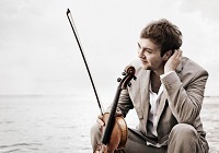 Mann mit beigem Anzug sitz am Wasser und hält eine Geige in der Hand.